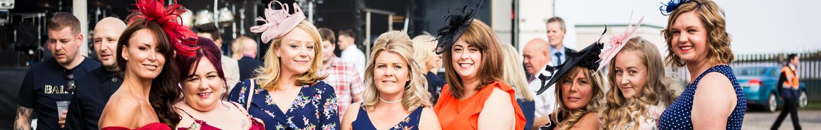A large group of ladies dressed up for the races pose for a picture in front of the stage.
